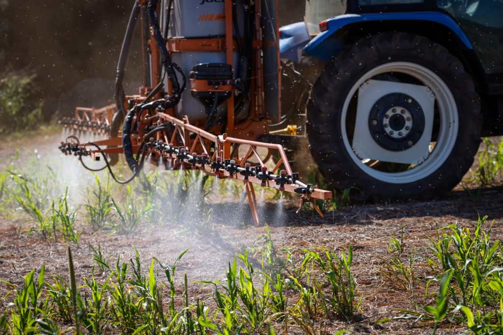 Fotografia colorida de uma máquina agrícola pulverizando defensores agrícolas no solo e nas plantas.